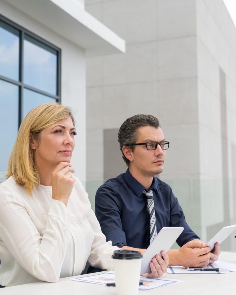 Pensive male and female colleagues working in outdoor cafe. Businesspeople sitting at table with building in background. Outdoor business workplace concept.